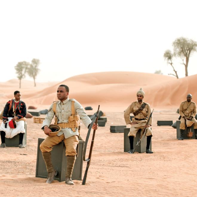 A photograph from the film Mimesis: African Soldier, with four men sat on grey blocks in a desert.