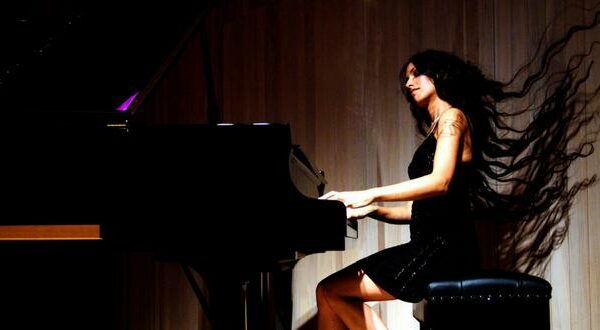 A photograph of Zoe Rahman performing, she plays the piano while her long hair flows in the wind