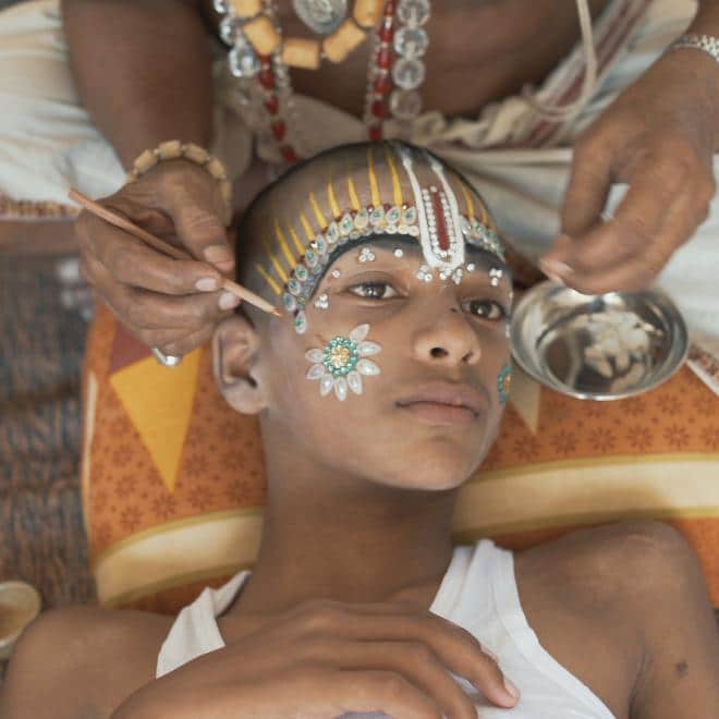 A photograph of an older woman decorating a young girl with jewels and makeup on her face as she's laying down on a pillow