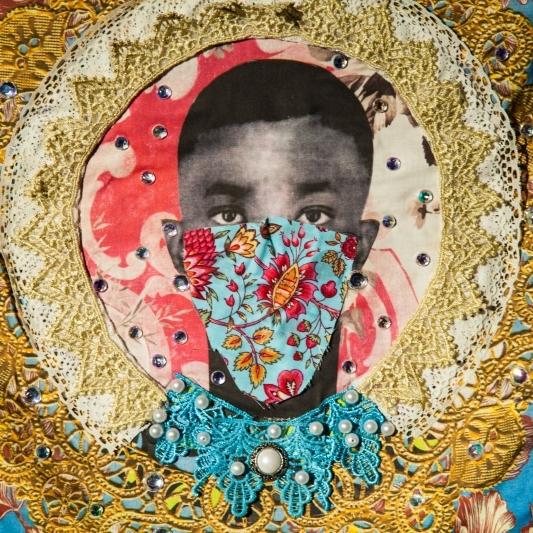 A photograph of a collage art piece depicting a cut out of a young boys head in black and white surrounded by yellow and blue lace and a patterned mask over his face