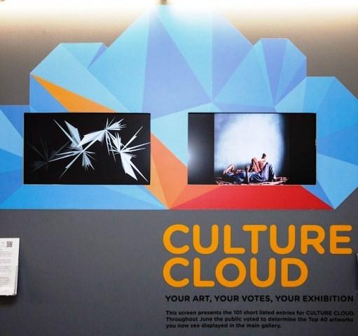 A photograph of an exhibition at the New Art Exchange, there is text on a wall which reads 'Culture Cloud, Your Art, Your Votes, Your Exhibition'