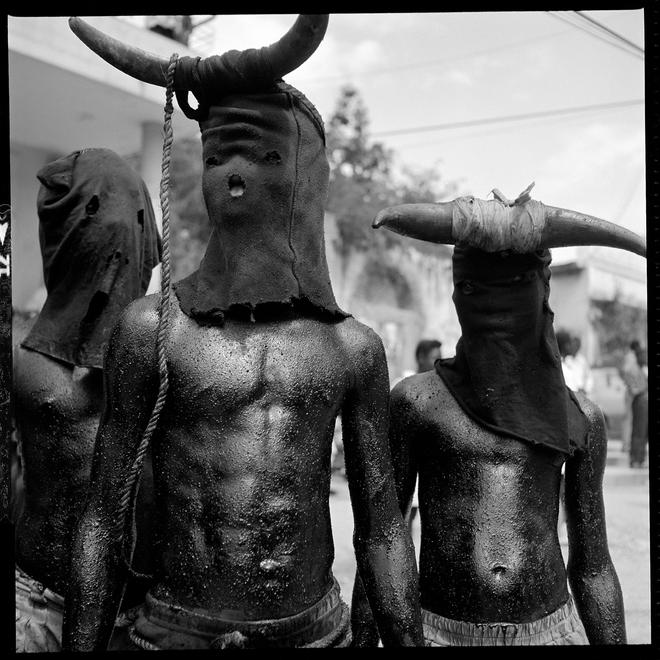 A photograph by Leah Gordon depicting three men with their shirts off wearing black cloth masks with horns attached to them which cover their whole face.