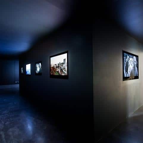 A photograph of an art exhibition at the New Art Exchange, there are tv's hung up on a wall casting a glow in a dimly lit room