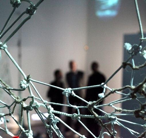 A photograph focused on pieces of string tied together to form a net, in the background there a three blurry silhouettes