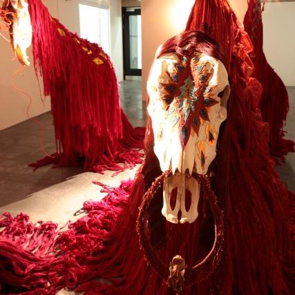 A photograph of a red woven art piece with an animal skull at the forefront of it, the piece resembles an animal and is hung from the ceiling
