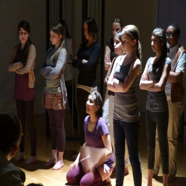 A photograph of a group of young people doing a theatre peformance, most are standing up with their arms crossed and one young girl is on the ground.