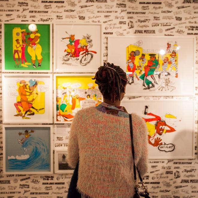 The back of a woman in a jumper, looking at brightly coloured art depicting cartoons, the art is hung up on a wall with a black and white typography pattern on it
