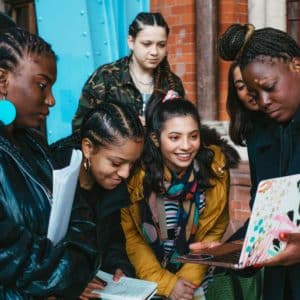 A group of young women from the Wondeherland collective. two of them holding books while all of them are staring to a laptop screen that a 5th woman is holding.