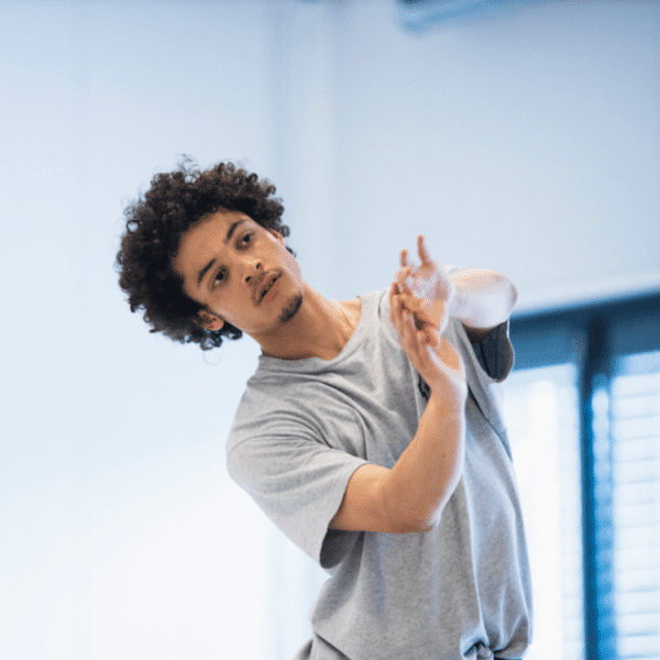 A picture of the artist Solomon Berrio-Allen. A mixed race male with curly hair, wearing a grey top, dancing in front of a grey background.
